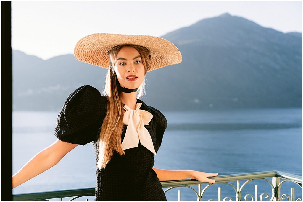 Woman wearing straw hat, black and white dress standing on balcony of hotel Tremezzo.